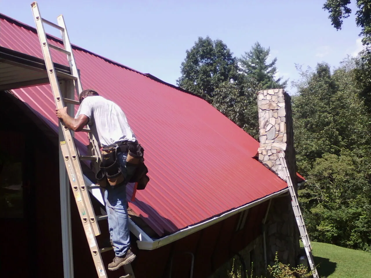 Roofer on a ladder beside a red metal roof and stone chimney — Soffit & Fascia Repair by Roofing Brothers in Mount Tremper